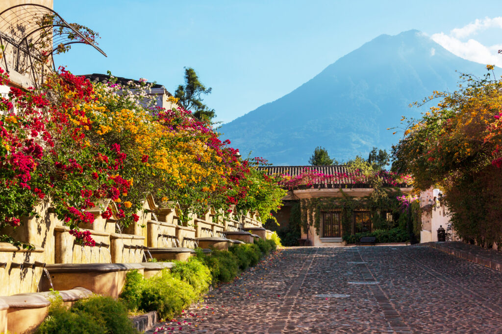 Calle empedrada de Antigua Guatemala con un arco colonial cubierto de buganvilia y el Volcán de Agua al fondo
